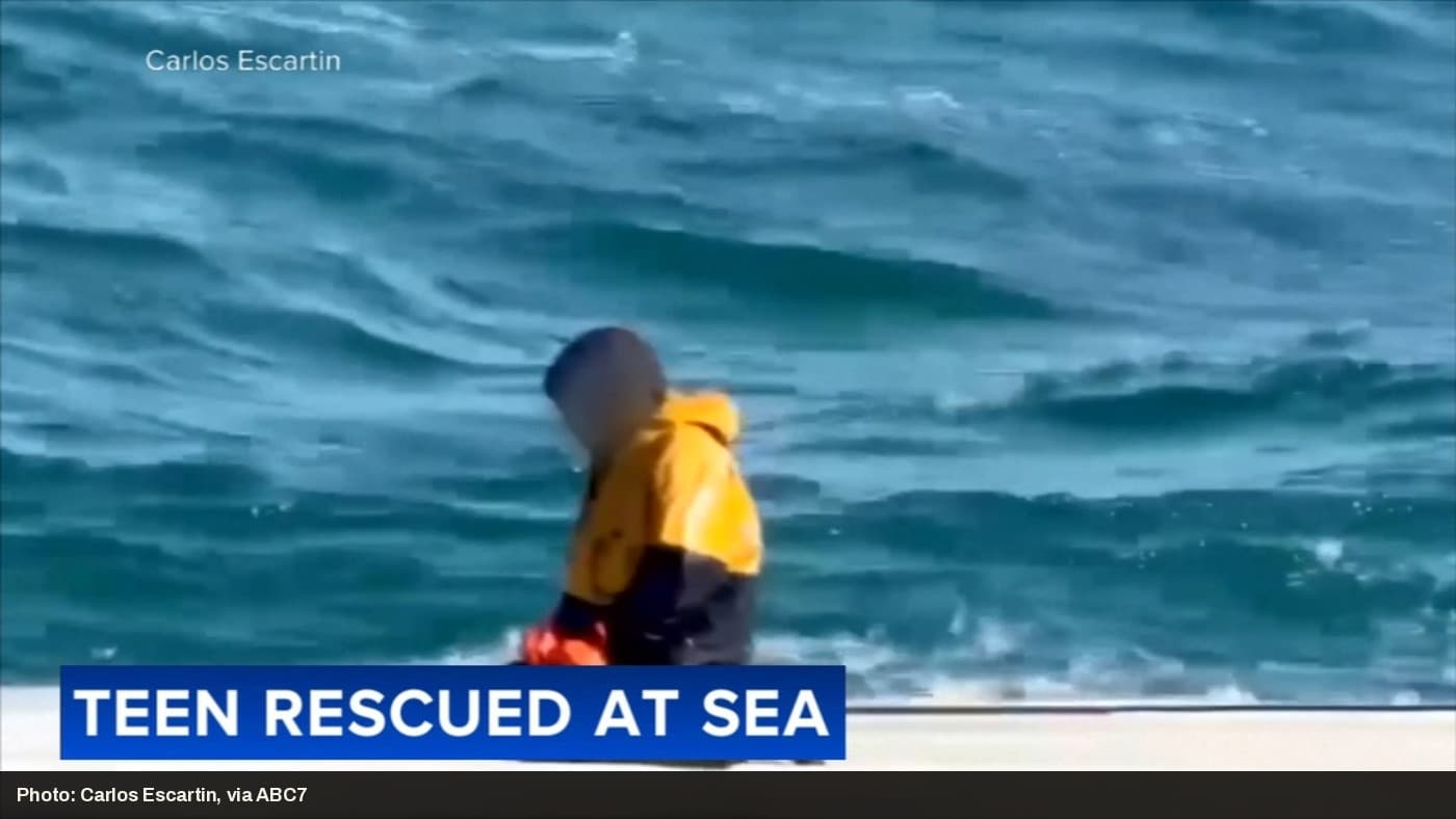 A rescued teen, face obscured, sits at the water's edge after being pulled from the Bering Sea.