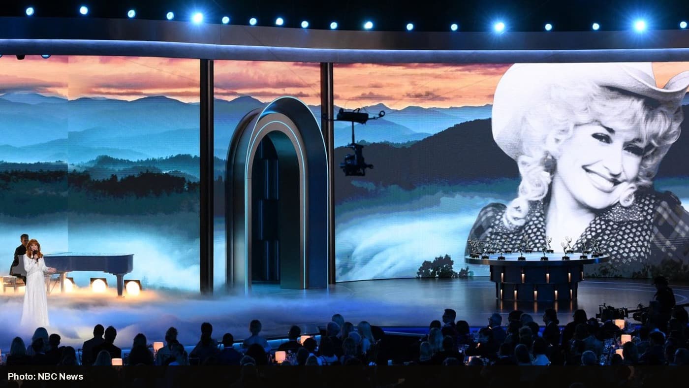 Reba McEntire performs at a piano on the Emmys stage beneath a giant black-and-white photo of Dolly Parton.