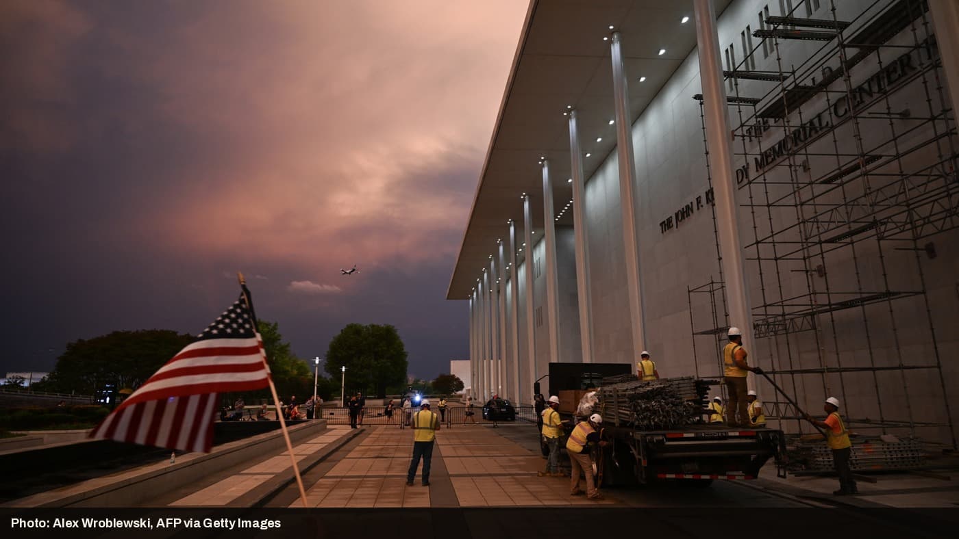 Construction crews work on scaffolding outside the Kennedy Center at dusk as an American flag waves in the foreground.
