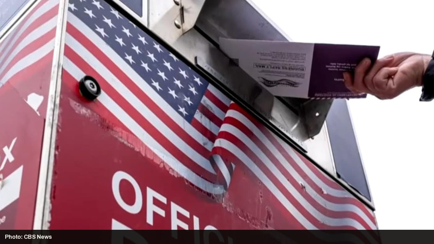 A hand places a mail-in ballot into an official USPS collection box.