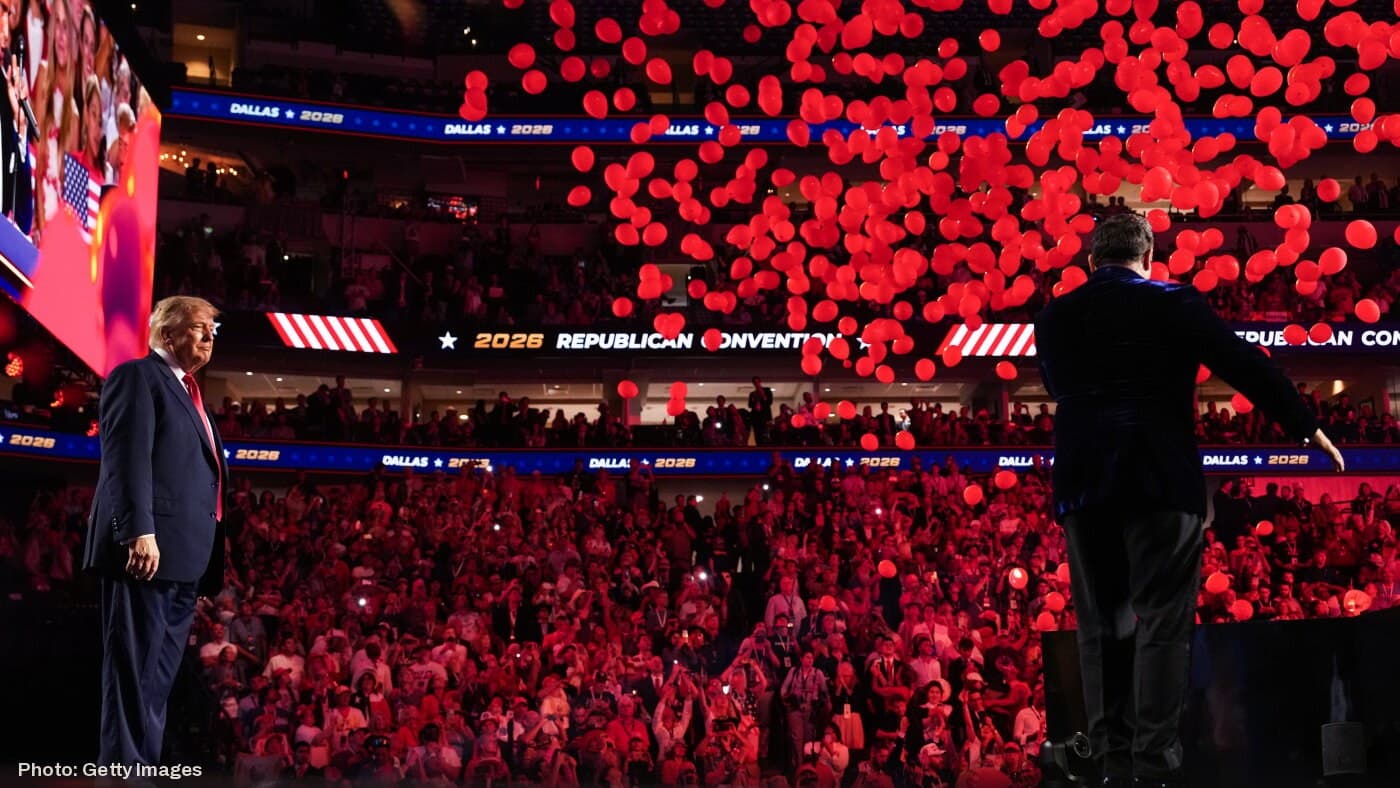 President Trump on stage at the 2026 Republican Midterm Convention in Dallas, Texas, as red balloons fall over the crowd.