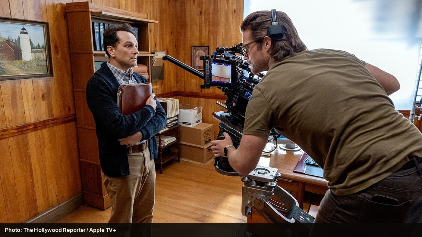 Behind-the-scenes shot of Widow's Bay being filmed, with an actor in a cardigan standing beside a film camera operator.