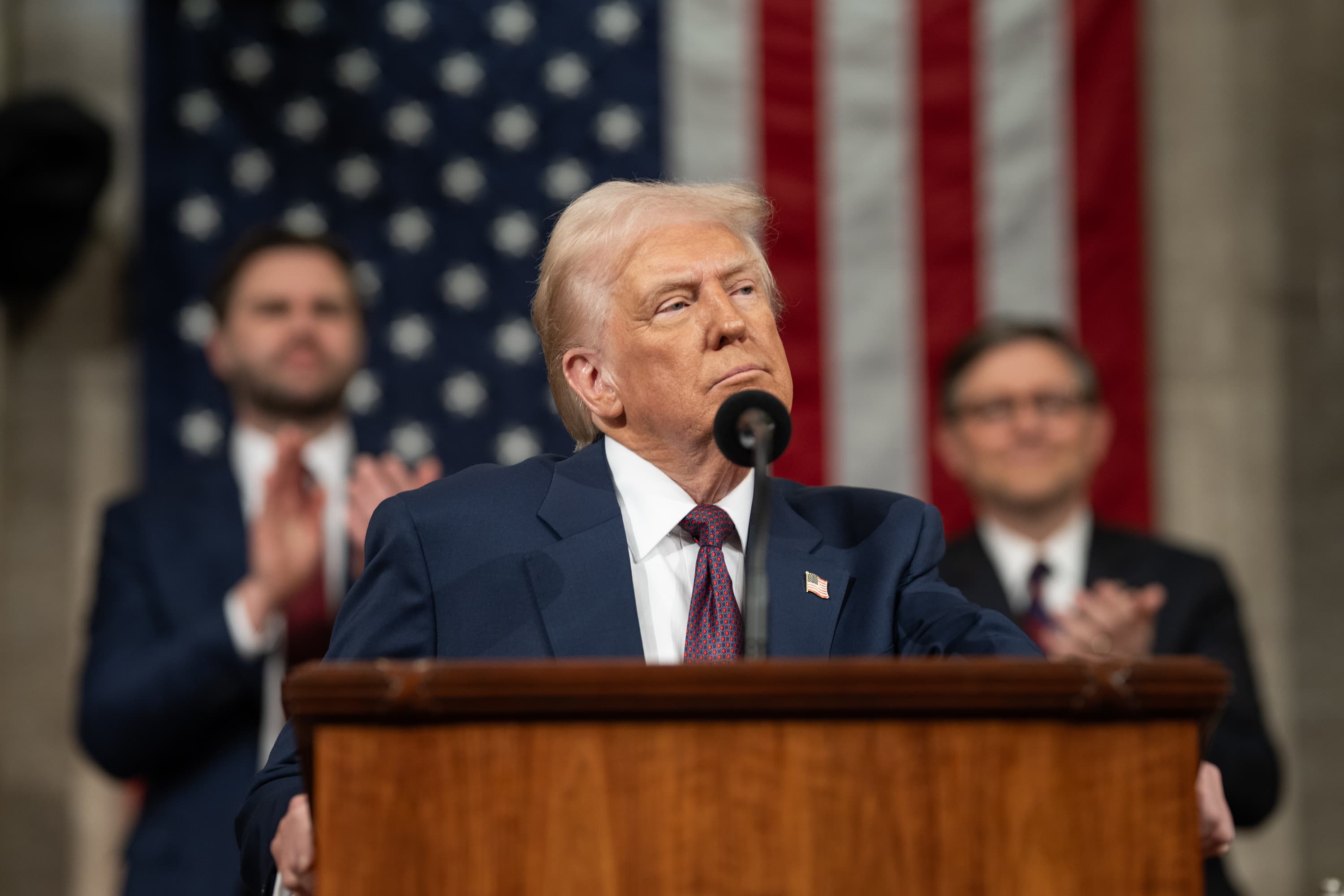 President Trump speaks at a podium with Vice President JD Vance and House Speaker Mike Johnson seated behind him