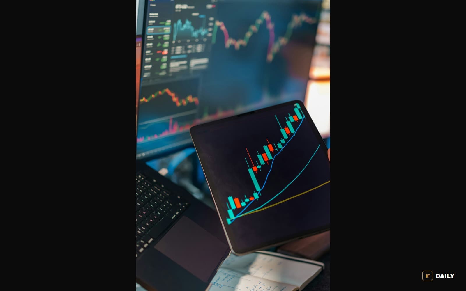A hand holds a tablet displaying a rising financial candlestick chart in front of computer monitors showing market data