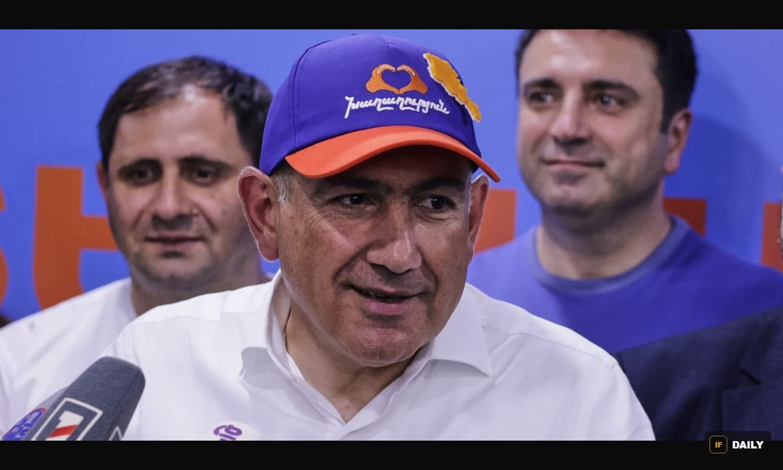 Nikol Pashinyan wearing a purple campaign cap, speaking to reporters with two men standing behind him