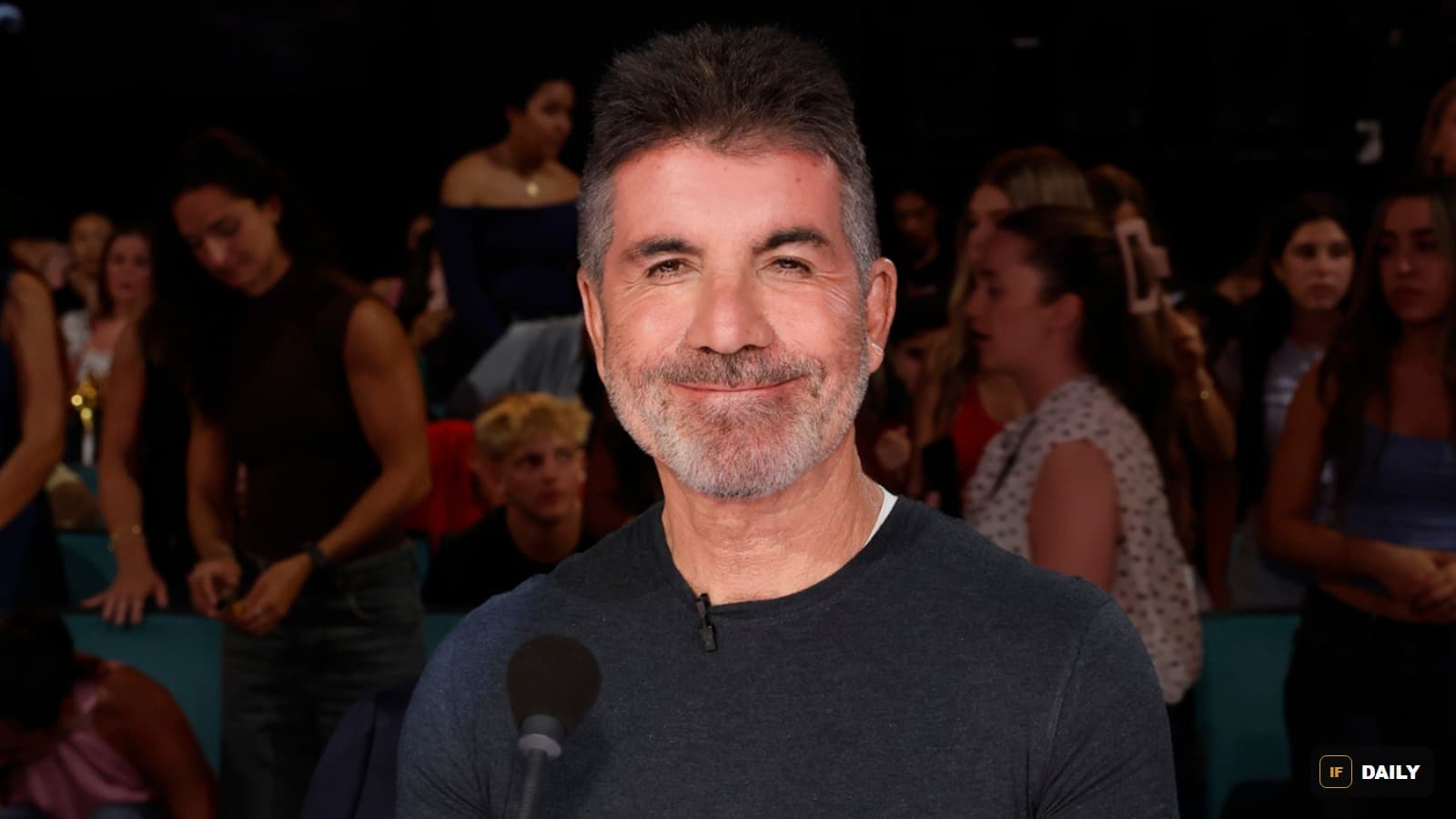 Simon Cowell smiling in a dark t-shirt in front of a studio audience