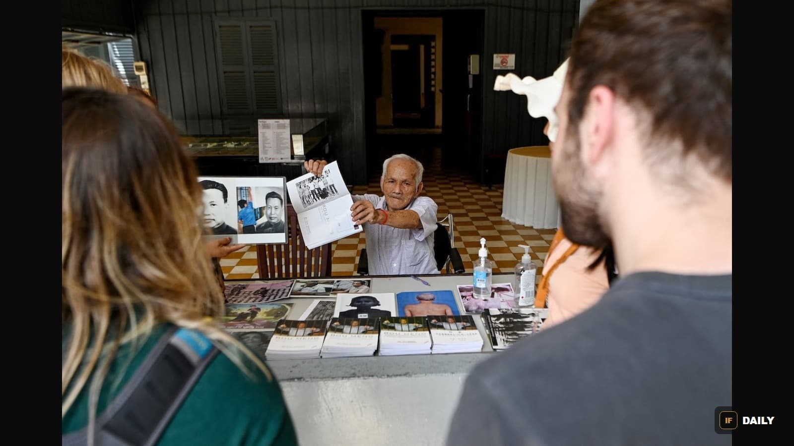 Bou Meng seated at a table showing a book of photographs to visitors at the Tuol Sleng Genocide Museum