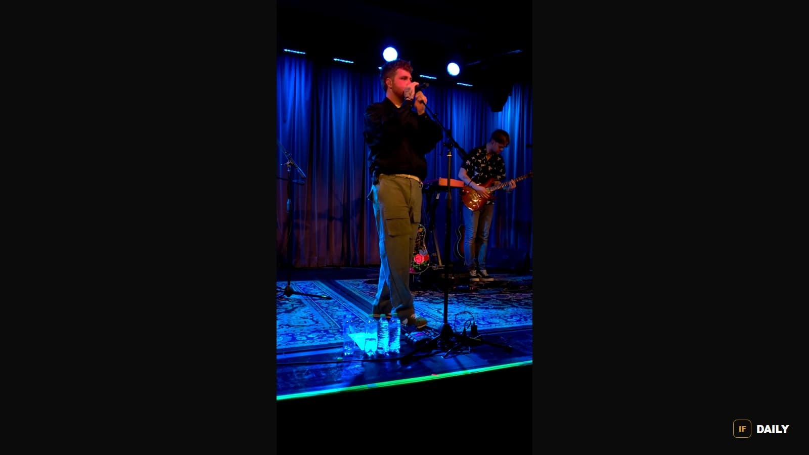 Alex Warren performing live and singing into a microphone on a small stage lit in blue