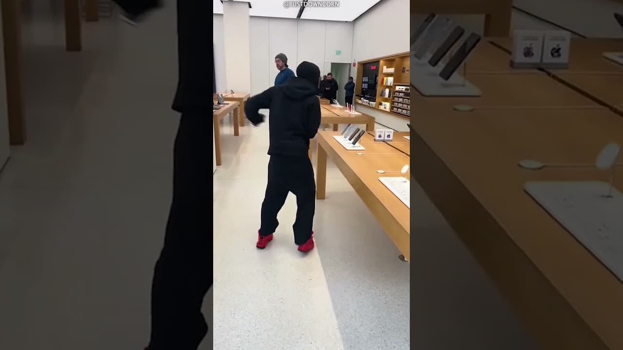 A masked man in a black hoodie and red sneakers grabs iPhones from a display table inside an Apple Store